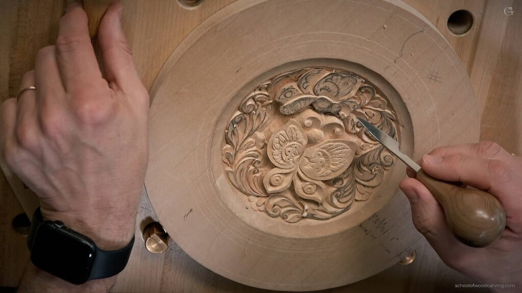A person's hands holding a carving knife and carefully carving a butterfly on a wooden plate. The process of creating the butterfly wall decoration involves intricate techniques and precision, as shown by the artist's focus and attention to detail. The use of Basswood and 10th century European style adds to the authenticity and beauty of the final carved piece.
