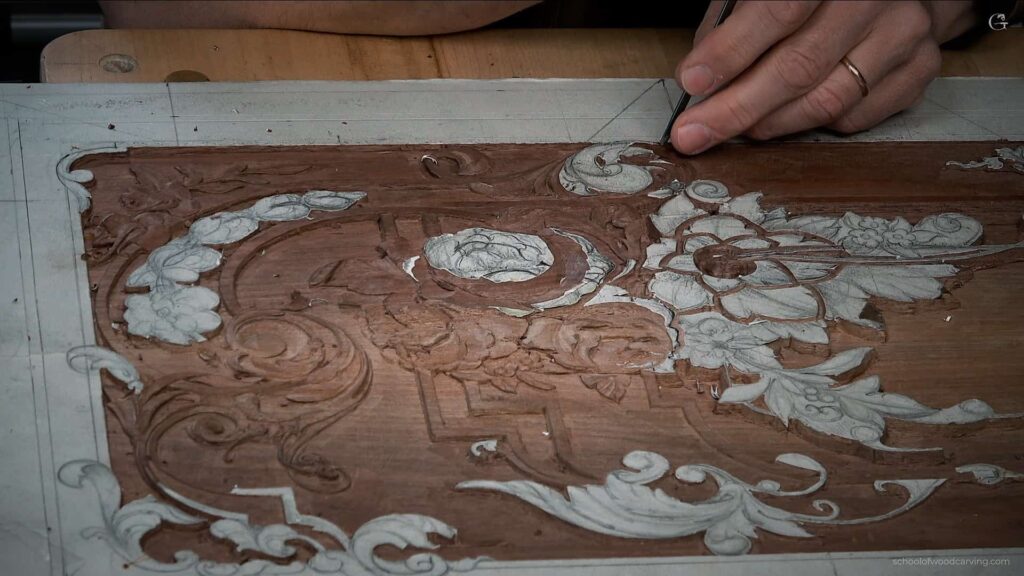 A wood carving artist using a razor blade to create intricate details on a wooden panel. Gouges and other carving tools are visible on the workbench. The artist's hands are focused and precise as they carve delicate curves and textures into the wood.
