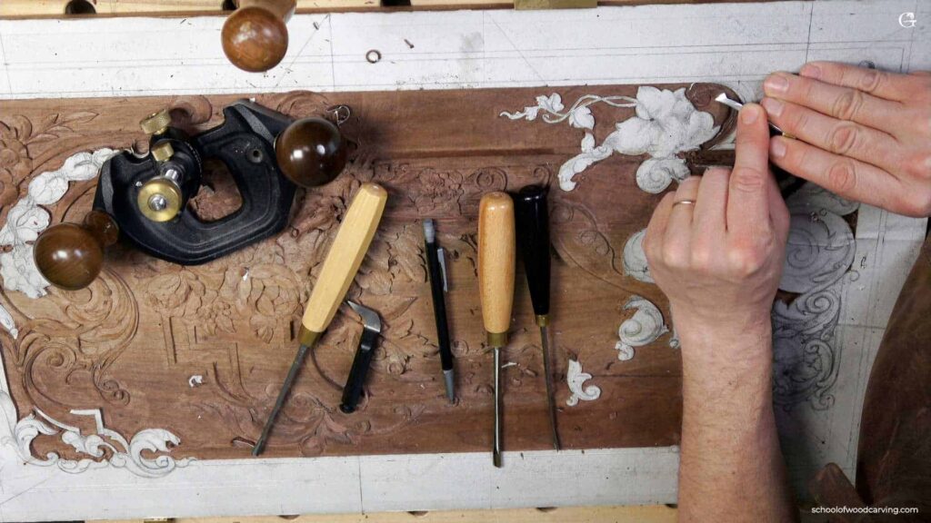 A person's hand carving a floral design on a wooden panel, surrounded by woodworking tools including chisels and a hand plane.