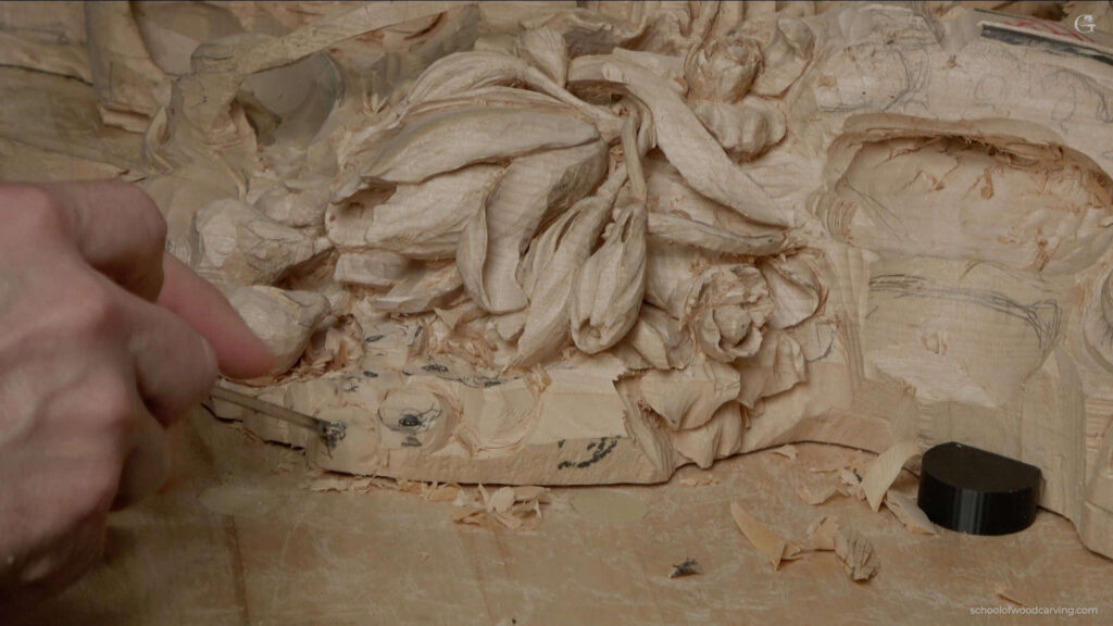 WOODCARVING LESSONS: A close-up photo shows Woodcarver Grabovetskiy using a chisel to carve beautiful flower shapes into a block of wood. You can see many wood shavings around the petals and leaves he is making. This picture shows his skill in woodcarving. If you want to learn how to carve wood like Grabovetskiy, join our woodcarving lessons or course. Our step-by-step tutorials will teach you how to create detailed patterns and improve your carving skills. - Wood Carving tutorials