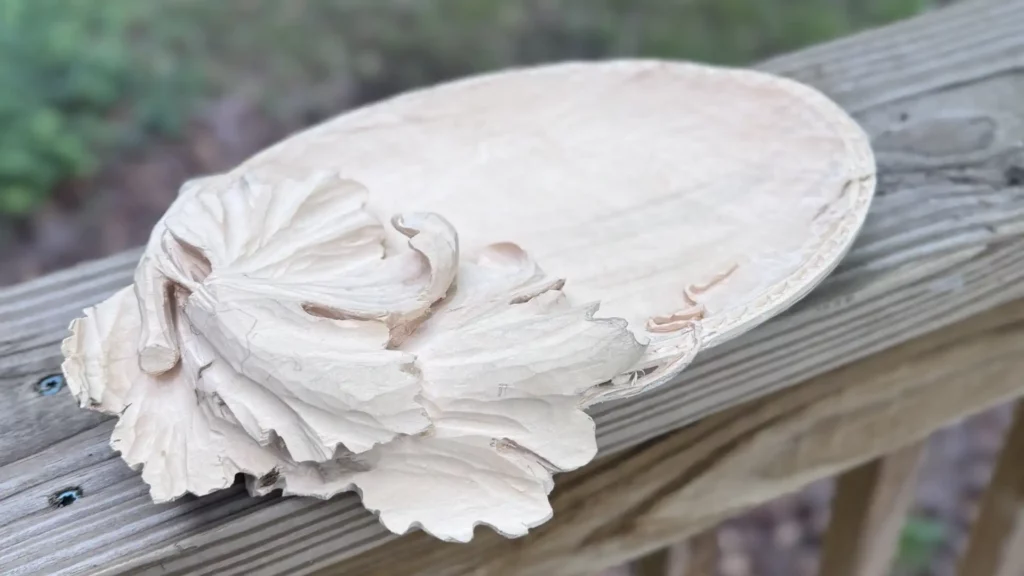 WOODCARVING LESSONS: A hand-carved wooden plate is shown in the picture. This plate is called a "Tray of Blessing." The tray has beautiful leaf and curled vine shapes carved on one side. The woodcarver, Grabovetskiy, made these detailed designs by hand. The tray sits on a wooden railing outdoors. In the background, you can see green plants and earth but they are blurry. The surface of the wood looks smooth with no finish yet. If you want to learn how to carve wood like this, join a woodcarving lesson or course by Woodcarver Grabovetskiy. In his tutorials, he teaches step-by-step how to make carved plates and other projects using simple carving techniques. [SEO Keywords: Woodcarving lessons, Beginner wood carving tutorial, Carved plate tutorial, Tray of Blessing design, Decorative wood carving patterns]. - Wood Carving tutorials