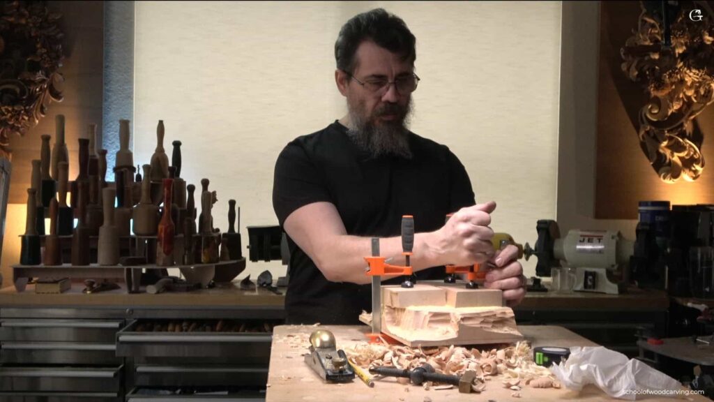 WOODCARVING LESSONS: Woodcarver Grabovetskiy uses clamps to hold wood on a workbench, with tools, shavings, and wood pieces in his woodworking studio. - Wood Carving tutorials